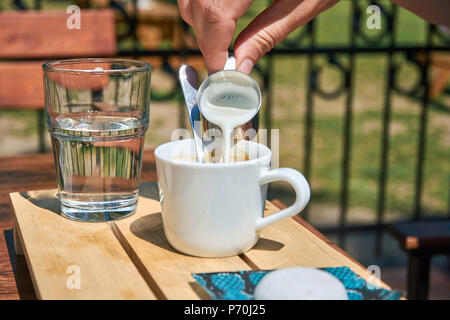 Une jeune femme dans un restaurant jardin verse du lait dans un café. Avec un verre d'eau et une table dans l'arrière-plan Banque D'Images