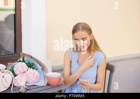 Portrait de femme à la mode la mode moderne message SMS par le smartphone en restaurant en plein air, d'attente, de rencontres. Bouquet de pivoines et de café sur le Banque D'Images