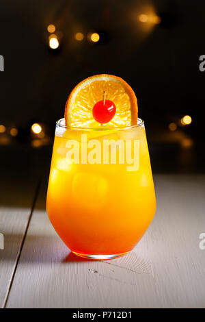 Tequila Sunrise avec une tranche d'orange et une cerise sur une table en bois blanc. Lumières de fête sur l'arrière-plan, la haute résolution Banque D'Images