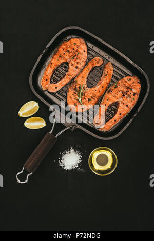 Vue de dessus les darnes de saumon sur le gril, sel, huile d'olive et citron vert tranches sur noir Banque D'Images