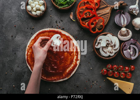 Cropped shot of woman putting tranche de champignons sur la pâte à pizza sur pizza sur table béton Banque D'Images