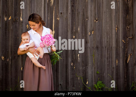 Une jeune femme brune dans une chemise blanche dans une jupe beige est tenant un petit bébé dans sa main et un beau bouquet de pivoines rose sur le background Banque D'Images