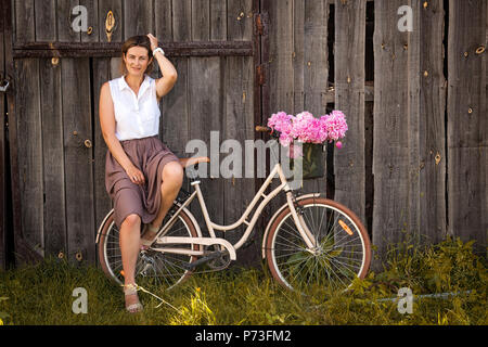 Une jeune femme brune dans une chemise blanche dans une jupe beige assis sur rond retro location de bouquet de pivoines rose dans un panier en osier sur la ba Banque D'Images