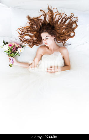 Vue de dessus bride wearing white dress sleeping in bed avec bouquet de fleurs Banque D'Images