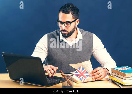 L'homme aux cheveux noirs l'apprentissage de l'anglais, le port de chandail gris veste et chemise blanche immaculée, travailler dur à l'office de bureau avec ordinateur portable avant mur noir Banque D'Images