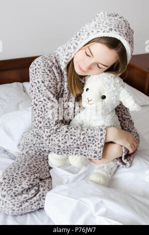 Jeune femme aux cheveux blonds assis sur le lit dans sa chambre à coucher dans une étreinte avec un animal en peluche jouet. Moutons préférés dans les mains de teen girl Banque D'Images