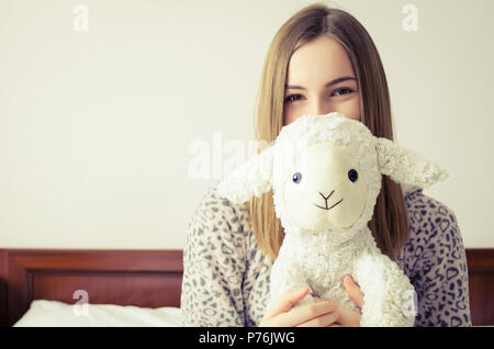 Jeune femme aux cheveux blonds assis sur le lit dans sa chambre à coucher dans une étreinte avec un animal en peluche jouet. Moutons blancs préférés dans les mains du t Banque D'Images