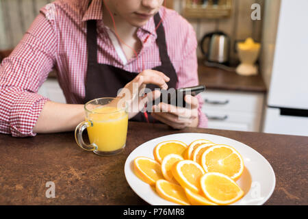 Jeune homme récolte des écouteurs assis à table de cuisine près de jus frais et de tranches d'oranges et de la navigation sur smartphone. Banque D'Images