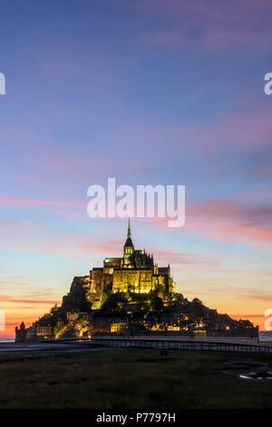 Vue sur le Mont-Saint-Michel tidal island en Normandie, France, allumé à la tombée de la jetée sur pilotis et pourpre des nuages dans le ciel. Banque D'Images