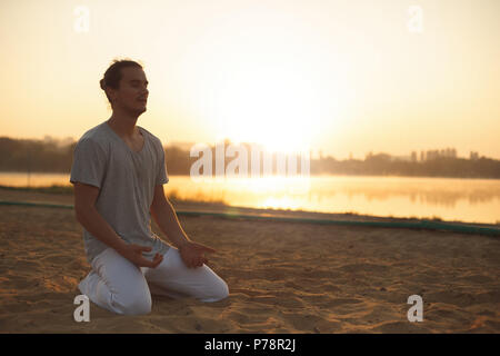 Jeune homme athlétique en pantalon blanc, posé sur la plage après l'entraînement du matin. La méditation, la réflexion, l'analyse de la vie. Coup de jeune homme sain en tenant Banque D'Images