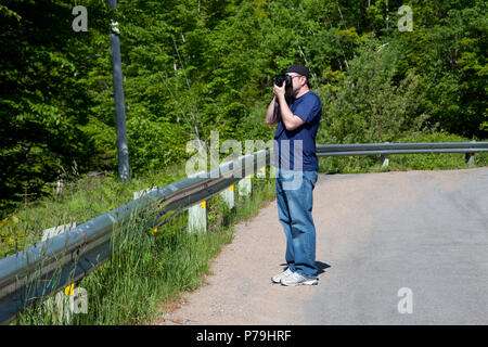 Un homme avec un appareil photo prend des photos à l'extérieur par les arbres feuillus sur l'accotement Banque D'Images