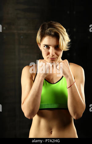 Close up portrait avant d'une jeune femme athlétique dans les vêtements de sport dans la salle de sport sur fond sombre, debout en position de boxe avec poings et mains, à la Banque D'Images