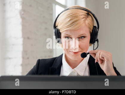 L'opérateur de centre d'appel concentré, femme blonde vêtue de noir veste et chemise blanche travaillant sur ordinateur portable par casque en bureau blanc Banque D'Images