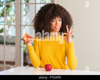 African American Woman choisir entre Apple et pizza slice faire ok signe avec les doigts, symbole excellent Banque D'Images