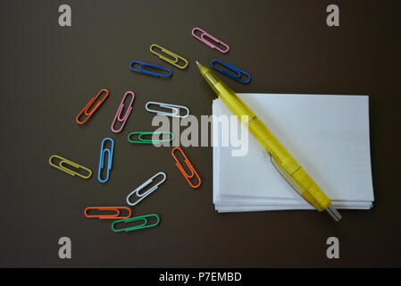 La couleur des trombones de couleurs différentes avec des feuilles de papier blanc pour l'enregistrement et un stylo jaune brun sur un fond mat. Banque D'Images