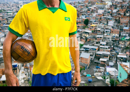 Joueur de football brésilien, qui se trouve dans l'équipe du drapeau Brésil vintage shirt couleur 1950 holding soccer ball en cuir brun traditionnel en face de favela Banque D'Images