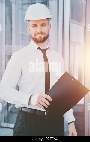 Un homme travaillant dans un casque blanc et chemise blanche avec une tablette dans ses mains contre l'arrière-plan d'un immeuble de bureaux en verre. Ingénieur. Planification des co Banque D'Images