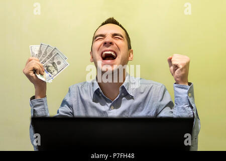 Jeune homme moderne excité avec son succès tout en travaillant avec son ordinateur portable. L'homme criant de joie, maintenant l'argent. 500 dollars dans la main Banque D'Images