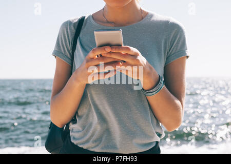 Jeune femme à la mode sms permanent sur le fond de mer et de soleil flare. Portrait de femme portant des T-shirt bleu et noir sac d'épaule u Banque D'Images