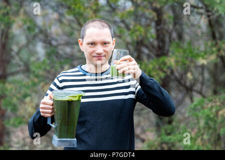 Jeune homme debout à l'extérieur, en plein air, holding plastic contenant du mélangeur, le verre avec smoothie vert fabriqué à partir de légumes verts, Banque D'Images