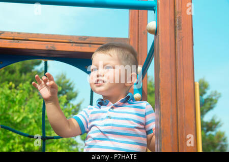 Caucasien mignon bébé garçon blond aux yeux bleus tout-petit de 2 ans se tient sur une aire de jeux dans le parc de la ville de sourire. Journée d'été lumineux vif feuillage vert B Banque D'Images