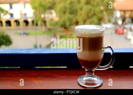 Cappuccino chaud dans une tasse transparente servis à la terrasse avec troubles de la place de la vieille ville de Cusco, Pérou en arrière-plan Banque D'Images