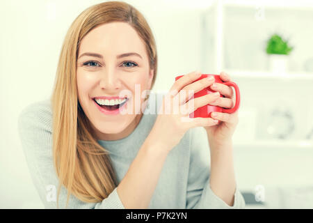 Une jolie jeune femme tenant une tasse rouge assis à une table dans un bureau blanc. Pause café. Banque D'Images