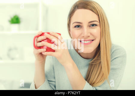 Une jolie jeune femme tenant une tasse rouge assis à une table dans un bureau blanc. Pause café. Banque D'Images