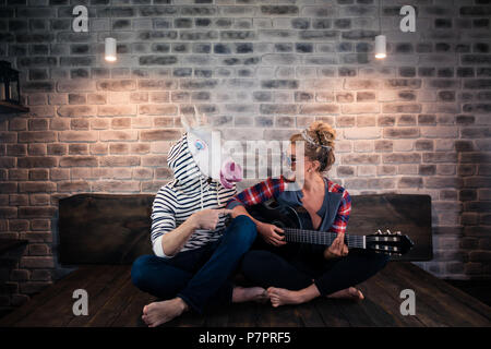 Jeune fille blonde à jouer de la musique à la guitare pour freaky homme en masque comique. Couple inhabituel passent du temps ensemble dans cet appartement élégant. Amie avec Unicorn Banque D'Images