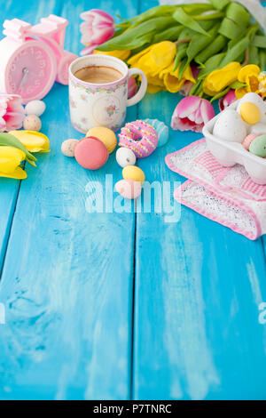 Les oeufs de pâques de diverses couleurs. Un réveil rose et une tasse de café, du café et un macaroni pour le petit-déjeuner. Fond en bois bleu et de tulipes au printemps. Une serviette rose. Espace libre pour le texte. Banque D'Images
