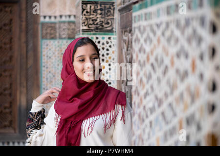 Portrait de femme musulmane smiling en costume traditionnel avec hijab rouge en face de mur décoré arabesque traditionnels Banque D'Images