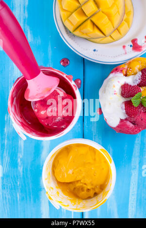 La crème glacée aux fruits à la mangue et les framboises dans un seau. Cuillère pour la crème glacée. Fond bleu et fleurs. Desserts sucrés Banque D'Images
