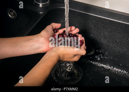 Groseilles rouges dans les mains du garçon de race blanche. Boy est maintenant venu sous l'baies humides de l'écoulement de l'eau du robinet et les laver. Évier noir élégant avec de l'eau Banque D'Images