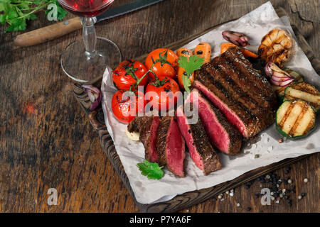 Tranches de bifteck grillé rares sur la planche à découper rustique avec set de légumes grillés sur table rustique foncé avec verre de vin rouge. Place pour le texte Banque D'Images