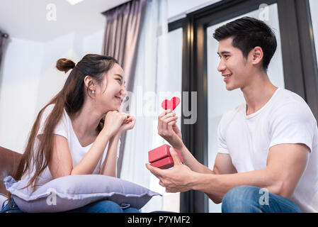 Man holding surprise fort et rouge coeur cadeau pour femme sur canapé à la maison dans la salle de séjour vous détendre et maison de concept. Happy sweet romantique de jeune couple Banque D'Images