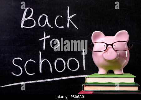 Pink piggy bank with glasses debout sur des manuels scolaires en face d'un tableau noir avec message retour à l'école. Mise au point nette sur la tirelire. Banque D'Images