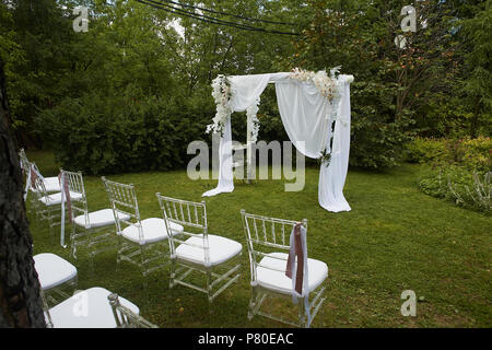 Cérémonie de mariage à l'air libre.chaises Transparent avec rubans voletant dans le parc ou la forêt. Arch décoré de fleurs et de mousseline blanche Banque D'Images