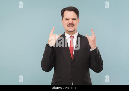 I'ts rock and roll, ouais ! Cool expressif happy rock signe. Piscine studio shot. isolé sur fond bleu. businessman avec costume noir, Banque D'Images