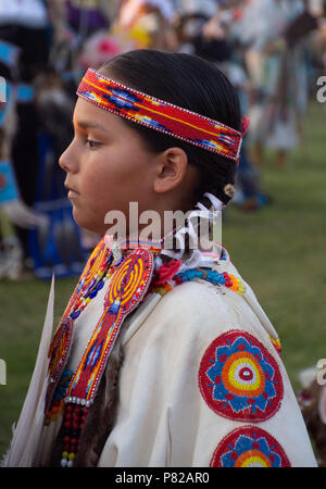 Close up of a Native American girl vêtue d'une robe blanche avec du rouge, bleu, orange, jaune et blanc avec médaillons et un bandeau perlé. Banque D'Images