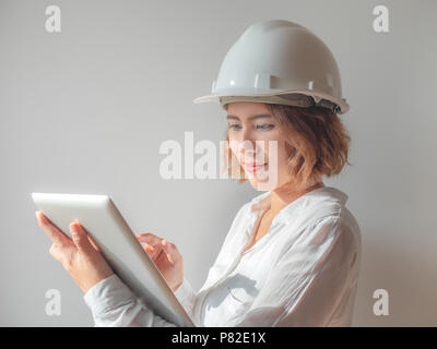 Belle asiatique ingénieur femme cheveux courts bouclés vêtu de blanc casque de sécurité et une chemise blanche avec de l'argent de travail comprimé et souriant. Femme au travail conc Banque D'Images