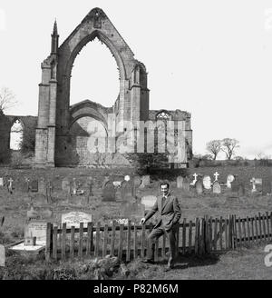 1959, historique, un homme debout par une petite clôture en bois par l'cementry qui se trouve en face des ruines de l'abbaye de Bolton dans Wharfedale, Yorkshire, Angleterre, Royaume-Uni. L'Augustin Prioy ly ruines à côté de la rivière Wharfe. Banque D'Images