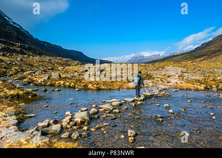 Walker crossing a' l'Allt Coire Dubh Mòr, entre Beinn Eighe & Liathach, Glen Torridon, région des Highlands, Ecosse, Royaume-Uni. Beinn Dearg dans la distance. Banque D'Images