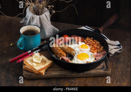 Petit déjeuner anglais avec saucisses, bacon, œufs frits, haricots, pain grillé et café sur table en bois Banque D'Images