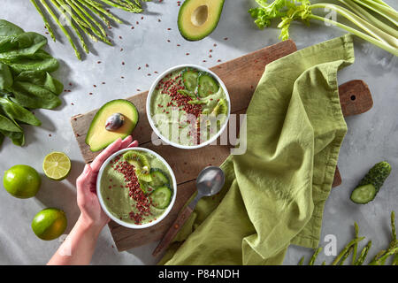 Les smoothies de légumes verts de désintoxication avec le concombre et les kiwis et les graines de lin dans une assiette sur une planche de bois. A woman's hand est titulaire d'une plaque. Vue d'en haut Banque D'Images