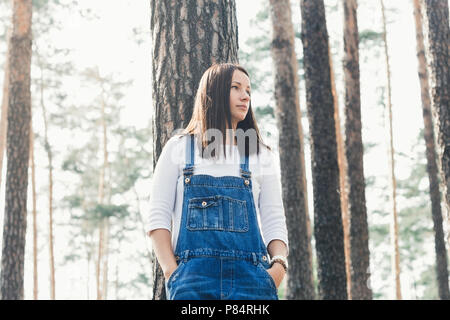 Belle jeune femme en jeans salopettes debout dans la forêt dans la soirée Banque D'Images