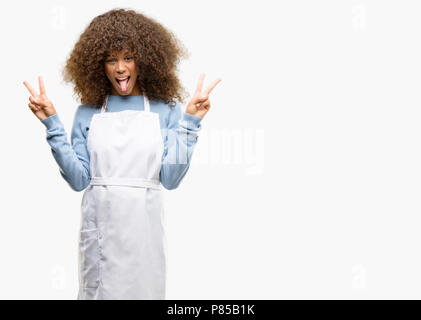 African American boutique femme portant un tablier, looking at camera montrant la langue et faire de la victoire avec les doigts Banque D'Images