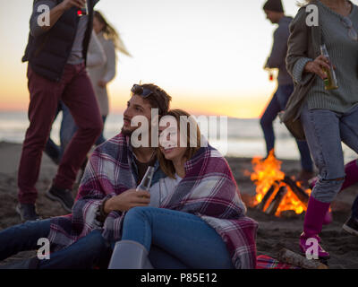 Jeune couple assis avec des amis autour de feu de camp sur la plage au coucher du soleil la consommation de bière Banque D'Images