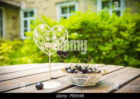 Cerises rouges frais de baies dans une assiette blanche sur la table en bois avec porte-bougie en forme de cœur à l'extérieur dans le jardin. Lieu Romantique. Banque D'Images