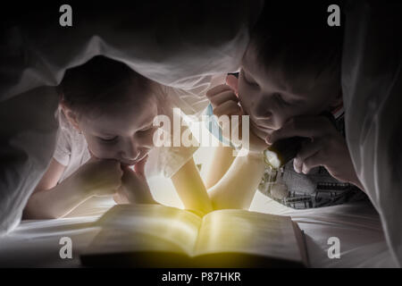 L'heure du coucher des enfants. Soeur et frère sont la lecture d'un livre sous une couverture avec une torche. Jeune garçon et belle fille s'amusant dans chambre d'enfant. Banque D'Images