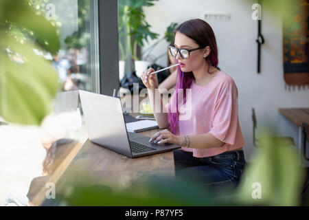 Vue latérale du young businesswoman sitting at table in coffee shop. Tasse de café sur la table et l'ordinateur portable. En arrière-plan mur blanc et fenêtre. Banque D'Images